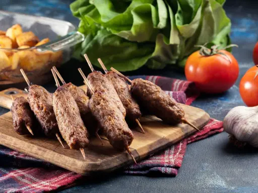 Aksamitne cevapi na drewnianej desce, obok pomidory, czosnek i pieczone ziemniaki.
