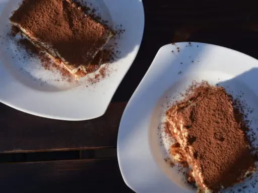 Dwa kawałki tiramisu posypane kakao na białych talerzach. Idealne do mocnej kawy, która podkreśli smak.