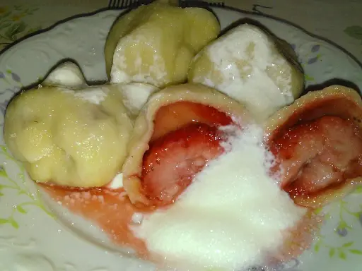Jak gotować pierogi mrożone z truskawkami? Na talerzu pyszne, gorące pierogi z owocami, polane śmietaną i cukrem.