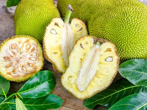 Cały i przekrojony chlebowiec (jackfruit) z widocznymi nasionami i liśćmi na drewnianym tle.