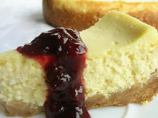 Kawałek kremowego sernika z polewą z ciemnych owoców na białym talerzu.