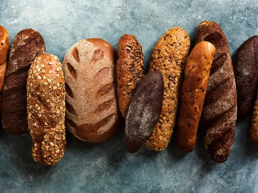 Różnorodne rodzaje chleba: bagietki, bochenki z ziarnami i ciemne pieczywo ułożone na niebieskim tle.