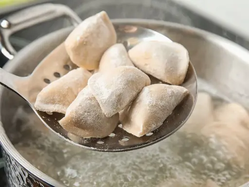 Mrożone pierogi na łyżce cedzakowej nad gotującą się wodą.