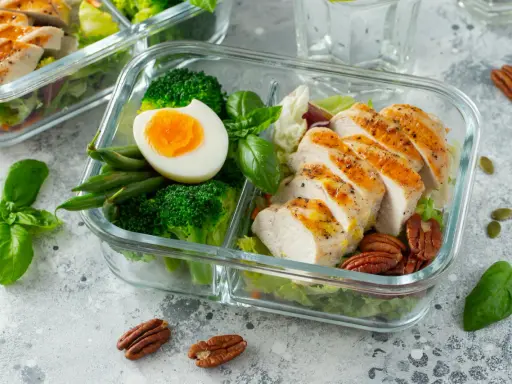 Szklane pojemniki z posiłkami: grillowany kurczak, brokuły, fasolka, jajko, orzechy i sałatka.