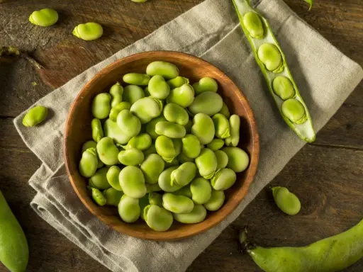 Miseczka pełna świeżych, zielonych fasolek fava, obok łuszczyn i pojedynczych ziaren na drewnianym blacie.
