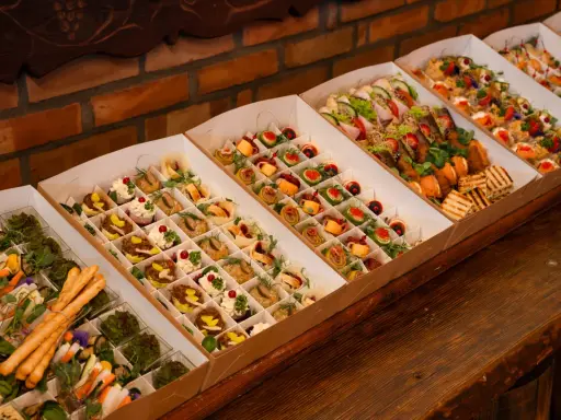 Elegancki catering do domu: pięknie ułożone przekąski w kartonowych pudełkach, od mini kanapek po rolki z łososiem i warzywami.