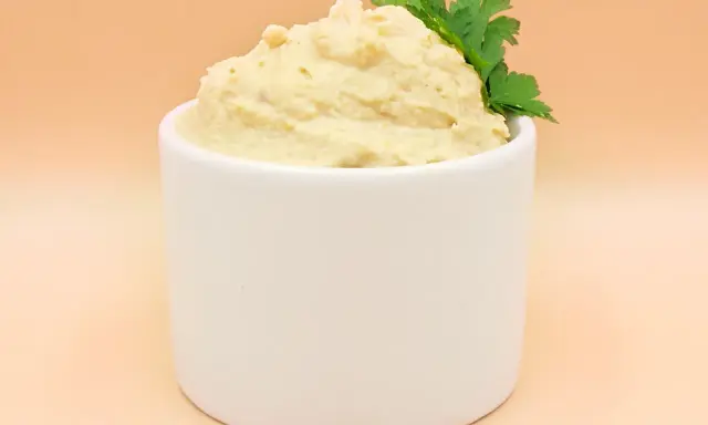 Kremowy hummus w białej miseczce, ozdobiony gałązką pietruszki.