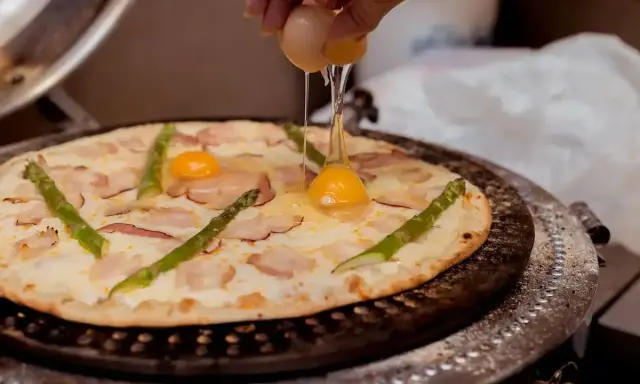 Pizza z jajkiem i szparagami, na którą wylewane jest surowe jajko.