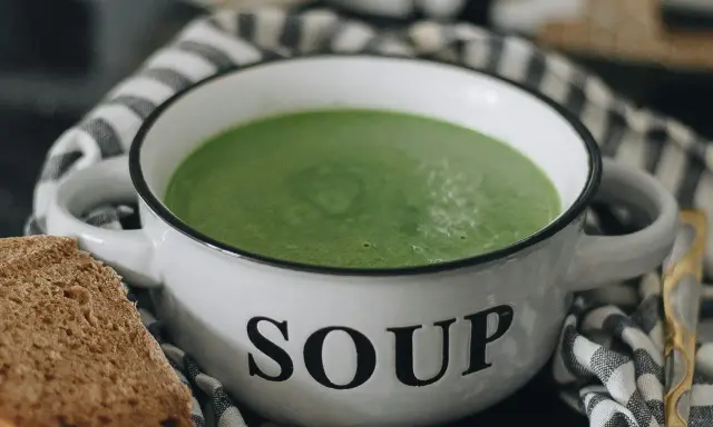 Zielony kremowy zupa w białej misce z napisem "SOUP", obok kromka chleba. Idealne rozwiązanie, jak mrozić zupy na później.