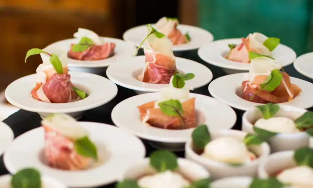 Eleganckie przekąski na białych talerzykach, zawinięte w szynkę parmeńską z listkami ziół. To właśnie catering, który zachwyca smakiem i wyglądem.