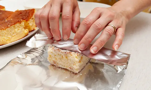 Ręce owijają kawałek ciasta w folię aluminiową. To prosty sposób, jak przechowywać szarlotkę, by dłużej zachowała świeżość.