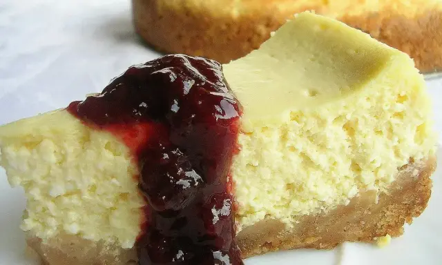Kawałek kremowego sernika z polewą z ciemnych owoców na białym talerzu.