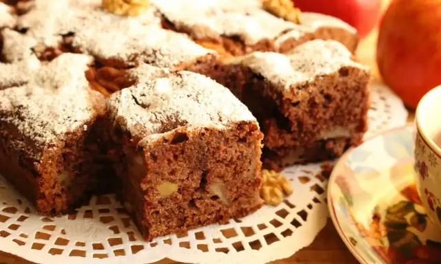 Kawałki aromatycznego ciasta cynamonowego z orzechami, posypane cukrem pudrem, obok jabłek i filiżanki herbaty.