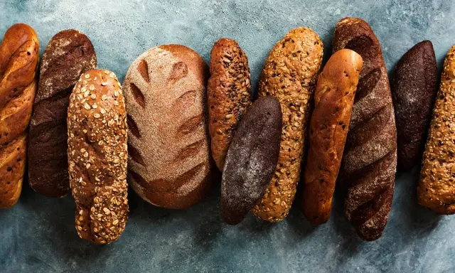 Różnorodne rodzaje chleba: bagietki, bochenki z ziarnami i ciemne pieczywo ułożone na niebieskim tle.