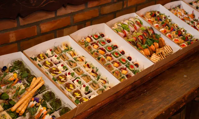 Elegancki catering do domu: pięknie ułożone przekąski w kartonowych pudełkach, od mini kanapek po rolki z łososiem i warzywami.