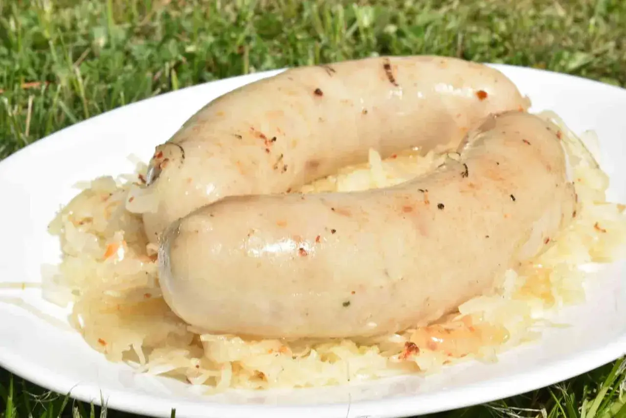 Dwie białe kiełbaski, idealnie przyrumienione na grillu, leżą na talerzu z kiszoną kapustą. Pyszna kiszka ziemniaczana z grilla w letnim otoczeniu.