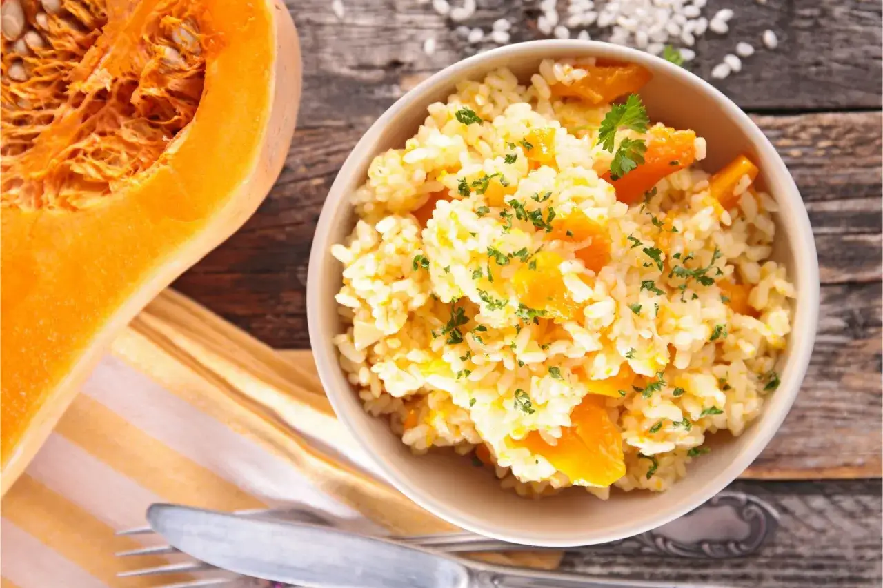 Pyszne risotto z dyni, idealne na jesienny obiad. Obok miski kawałek świeżej dyni i sztućce.
