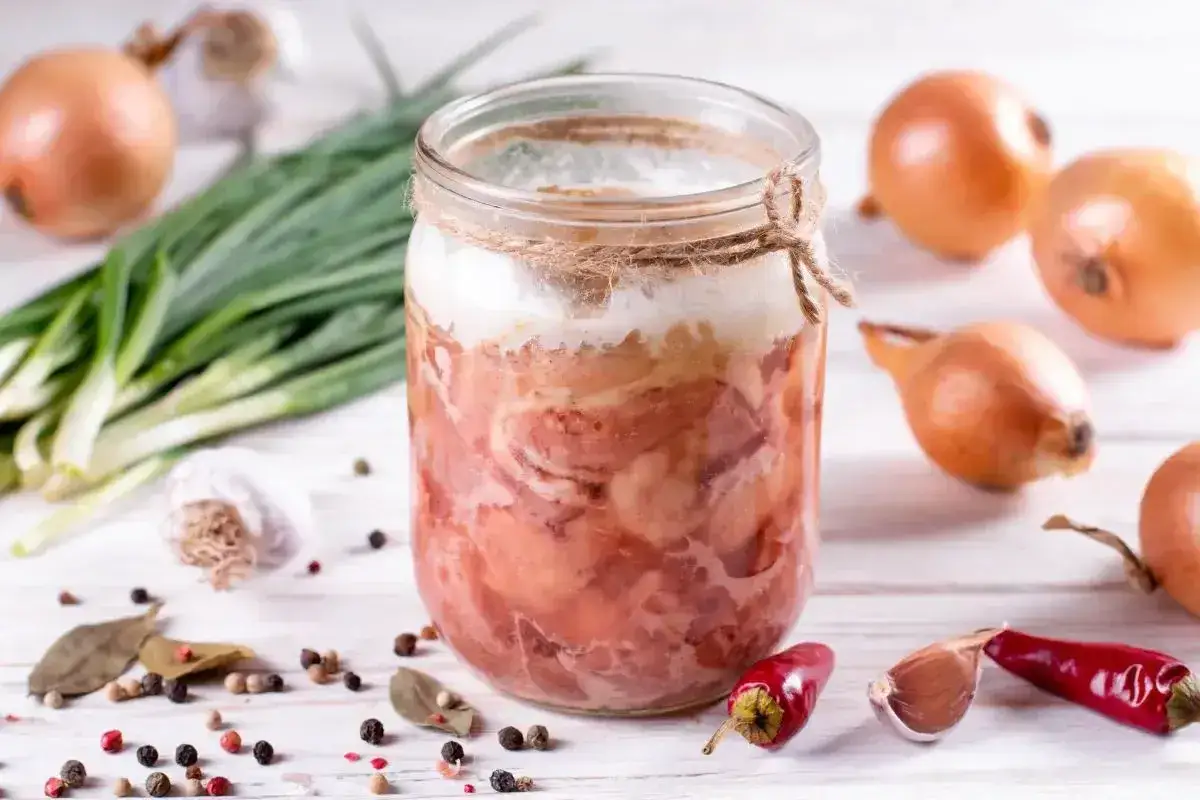 Słoik z domową kiełbasą w galaretce, otoczony cebulą, czosnkiem, pieprzem i papryczkami chili. Idealny przepis na to, jak zrobić kiełbasę w słoiku z galaretką.