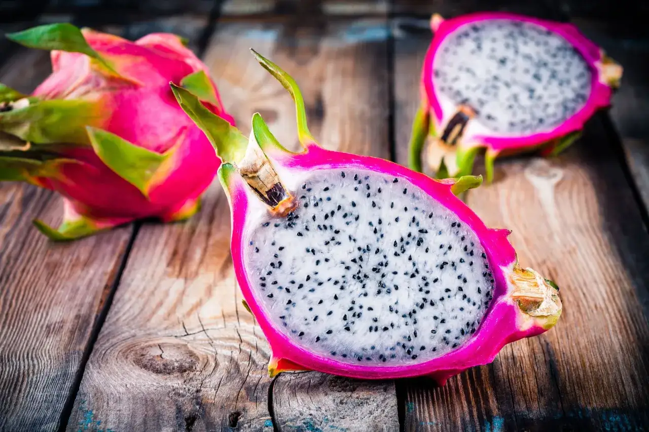 Dwie połówki i cała pitahaya czerwona z białym miąższem i czarnymi pestkami na drewnianym blacie.