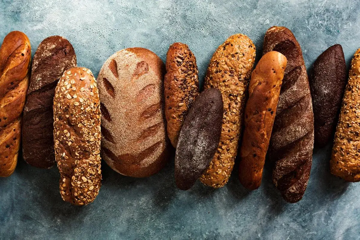 Różnorodne rodzaje chleba: bagietki, bochenki z ziarnami, ciemne i jasne pieczywo ułożone obok siebie.