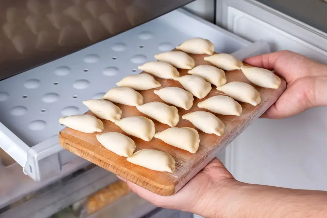 Ręce wkładają deskę z surowymi pierogami do zamrażarki. To prosty sposób, jak mrozić pierogi, by cieszyć się nimi później.