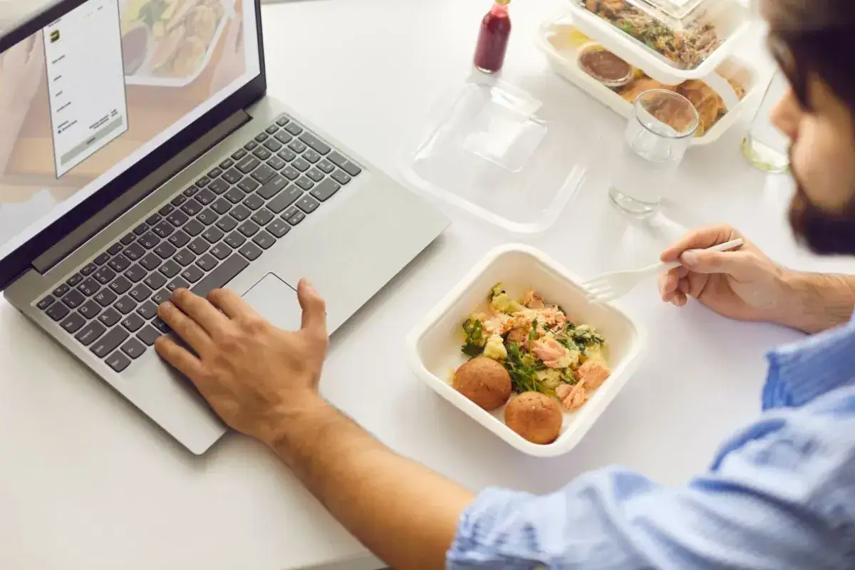 Mężczyzna pracuje przy laptopie, jedząc posiłek z cateringu. Analizuje, ile można zarobić na cateringu, planując biznes.