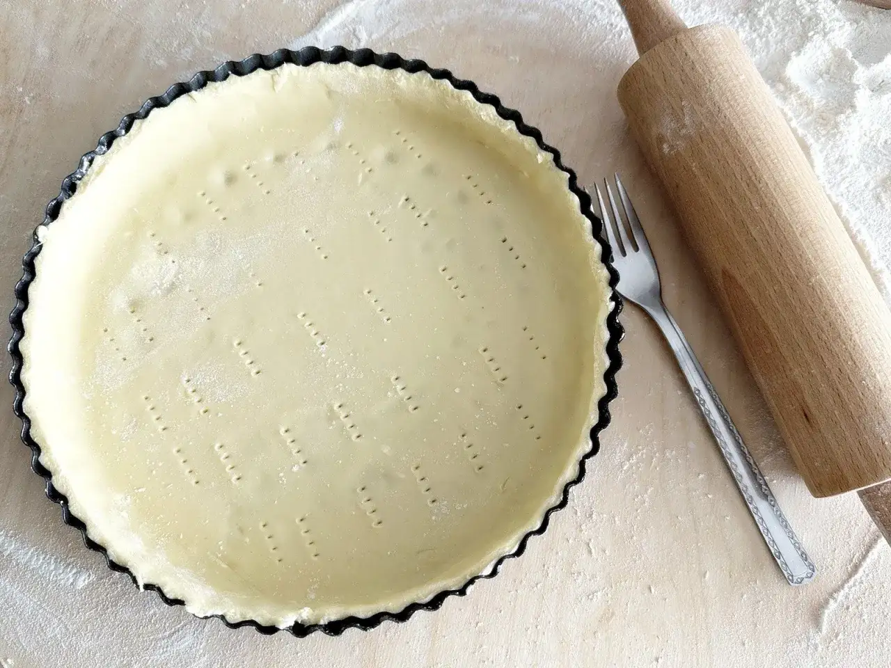 Przygotowania do pieczenia kruchego ciasta. Surowe ciasto w formie, wałek i widelec na blacie posypanym mąką.
