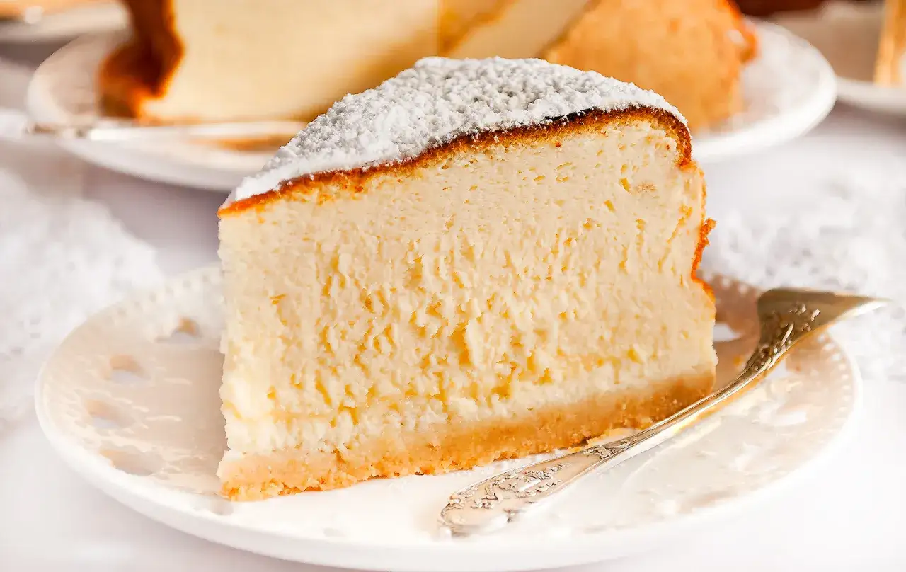 Puszysty sernik ze skyru i budyniu, oprószony cukrem pudrem, na białym talerzyku z ozdobnym widelczykiem.