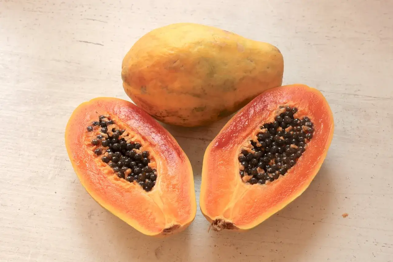 Dojrzała papaya, jedna w całości, dwie połówki z czarnymi nasionami, na jasnym tle.