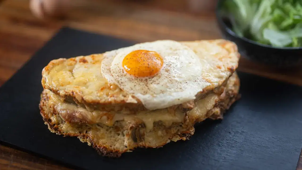 Pyszny croque madame z rozpływającym się serem, szynką i jajkiem sadzonym na wierzchu, podany na czarnej desce.