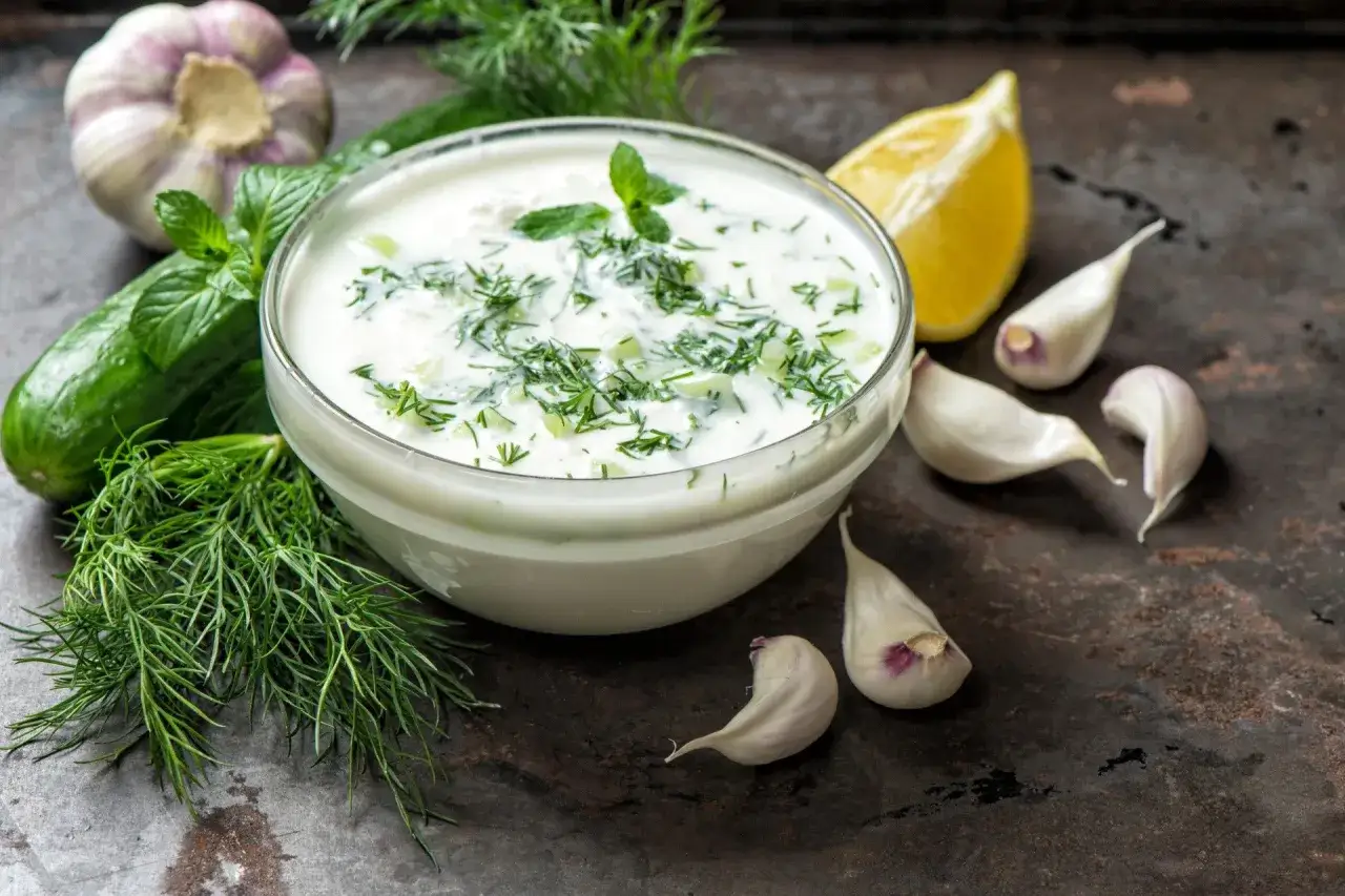 Pyszny sos czosnkowy jak z restauracji, z dodatkiem świeżego ogórka, koperku i mięty. Obok czosnek, cytryna i pęczek koperku.