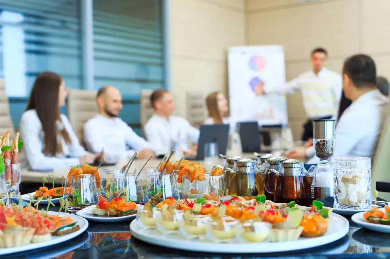 Elegancki catering na spotkaniu biznesowym: ludzie słuchają prezentacji, a na stole czekają przekąski.