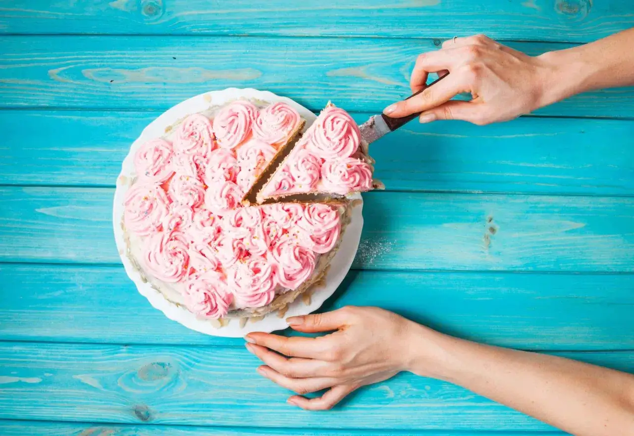 Ręce kroją różowy tort ozdobiony kremem i posypką na niebieskim tle.