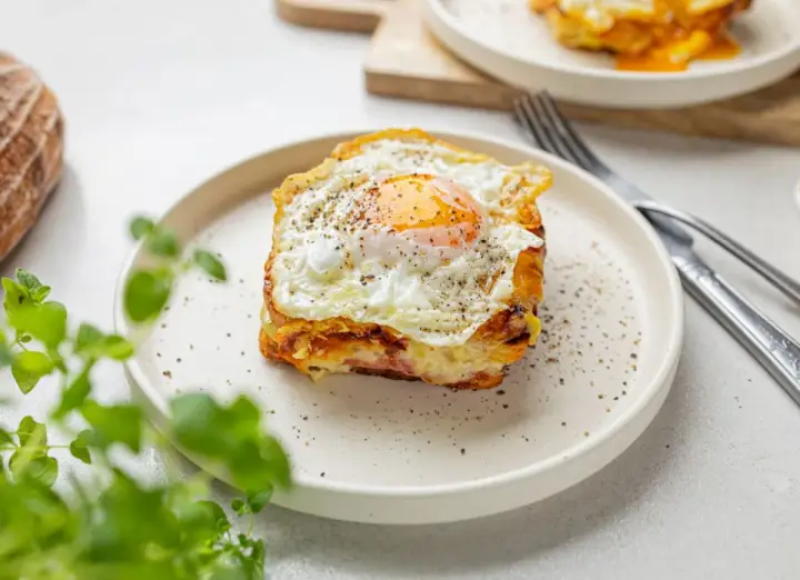 Pyszny croque madame na talerzu, z jajkiem sadzonym na wierzchu i pieprzem. Obok widoczny widelec i nóż.