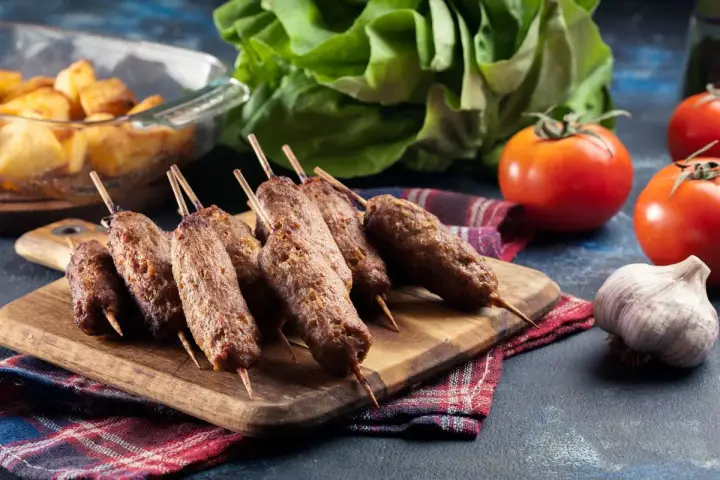 Aksamitne cevapi na drewnianej desce, obok pomidory, czosnek i pieczone ziemniaki.